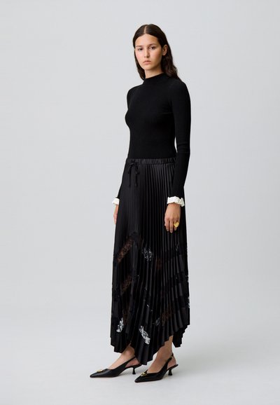 Haut noir à manches longues avec des côtes, associé à une jupe plissée noire avec des accents en dentelle. Le mannequin porte des escarpins noirs à bout pointu et une bague en or.