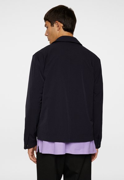 Giacca blu navy realizzata in tessuto morbido, con colletto, maniche lunghe e polsini con bottoni, indossata su una camicia di un leggero colore viola.