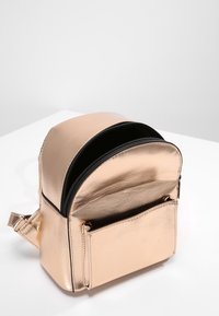 Metallinvärinen kultainen reppu, jossa on pyöreä muoto, vetoketjulukko ja etutasku. Silkkinen pinta yhdellä säädettävällä hihnalla.