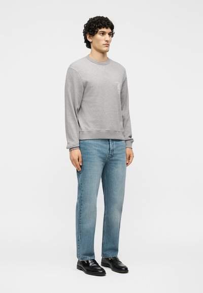 Νέος άνδρας που στέκεται, φοράει ανοιχτό γκρι φούτερ, ανοιχτά μπλε τζιν και μαύρα δερμάτινα μοκασίνια σε λευκό φόντο.