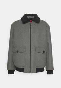 Grå ullbombarjacka med svart fårskinnskrage, frontdragkedja, två fickor med knappflik och svarta ribbade muddarna och nederkant.