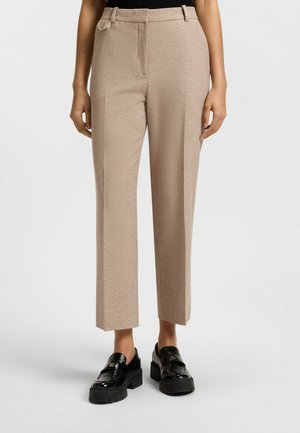 Pantaloni beige a gamba ampia realizzati in un tessuto texturizzato, con tasche laterali e una finitura lucida, abbinati a mocassini neri dal design chunky.