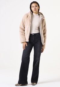 Chaqueta de piel sintética en beige claro con cremallera; combinada con jeans negros de campana. El conjunto incluye una parte superior de color crema con relieve y zapatos metálicos.