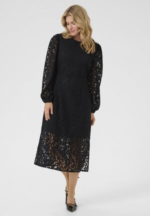 Black lace dress with long, puffed sleeves and a mid-length cut. Sheer floral pattern with solid underlay. Worn with pointed black heels.
