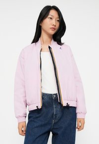 Jeune femme portant une veste bomber rose clair sur un débardeur blanc et un jean taille haute bleu, se tenant devant un fond blanc.