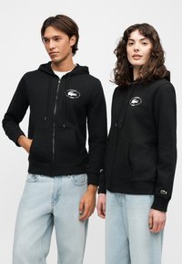 Homme et femme portant des sweats à capuche zippés noirs avec logos Lacoste, assortis à des jeans bleu clair, debout devant un fond uni.