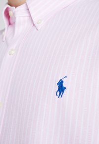 Camicia a righe rosa chiaro con bottoni, realizzata in un tessuto texture. Presenta un logo blu di un giocatore di polo sul lato sinistro del petto e dettagli dei bottoni bianchi.