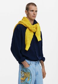 Marineblaue gerippte Button-Up-Bluse, gelber gestrickter Schal um den Hals drapiert, hellblaue Jeans mit Tigergrafik auf der Tasche.