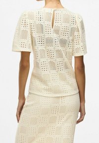 Bakifrån av en person som bär en krämfärgad kortärmad topp och en matchande kjol med ett rutigt virkade mönster och en liten nyckelhålöppning bak.