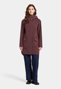 Donker burgundy waterdichte parka met capuchon, voorzijde met rits en drukknopen. Voorzien van twee voorkruiszakken en een rechte snit, gecombineerd met donkere spijkerbroeken.