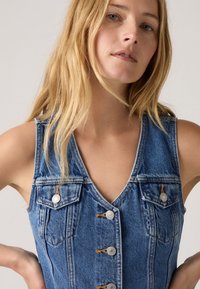 Gilet en denim avec un décolleté en V, de couleur bleu clair, doté de deux poches poitrine, de boutons argentés et d'une texture légèrement déchirée.
