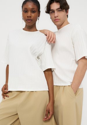 Deux mannequins, l'un à la peau foncée et l'autre à la peau claire, portent des hauts blancs côtelés à manches courtes et des pantalons larges beiges, se tenant près l'un de l'autre.