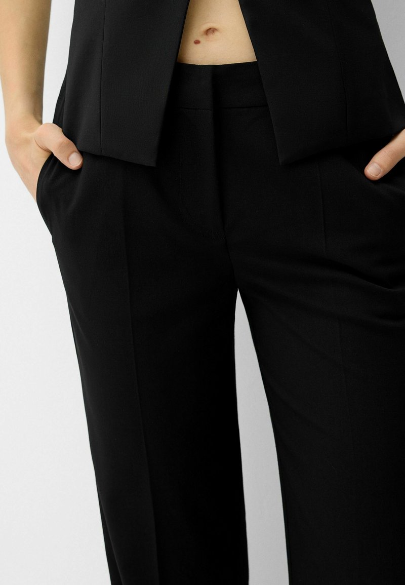 Pantalones negros a medida, con una tela suave, pierna ancha y bolsillos laterales. La cinturilla se sitúa justo por debajo de la cintura, con un acabado limpio.