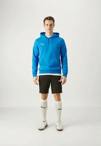 Modrá mikina s klokankou, čierne šortky, biele ponožky po kolená a biele tenisky. Na oblečení sa nachádza logo Puma. Jednoduchý športový dizajn.