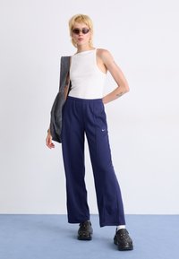 Witte geribbelde tanktop, navyblauwe losvallende joggingbroek met zijstrepen, zwarte klompen en een grijze jas over de schouder gedrapeerd.