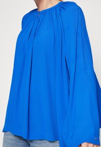 Blusa blu con design a pieghe, scollo rotondo e maniche lunghe e larghe. Realizzata in tessuto leggero, presenta un piccolo logo sul bordo.