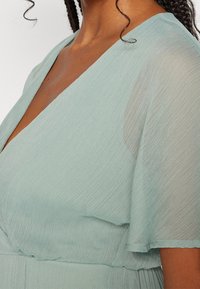 Robe enveloppante bleu clair avec un décolleté en V et des manches volantes. Fabriquée en tissu texturé et transparent, elle présente un corsage ajusté et une jupe fluide.