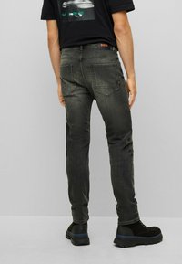 Dark gray denim jeans with a fitted design, featuring back pockets and a logo patch on the waistband, paired with black boots.