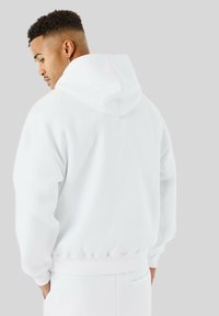 Man met kort krullend haar, gekleed in een eenvoudige witte hoodie en witte broek, met zijn rug naar de camera toe tegen een lichtgrijze achtergrond.