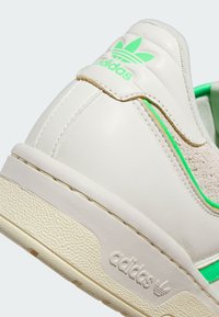Bílé kožené tenisky s zelenými akcenty, texturovaná zadní část a gumová podrážka s výrazným logem Adidas. Zaoblený design.