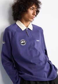 Lilla langermet polo skjorte med en kremfarget krage, logo-patcher og en avslappet passform. Glatt stoff med en subtil glans og ingen mønstre.