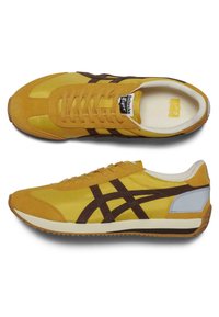 Žluté tenisky s hnědými akcenty, z nubuku a nylonu, zaoblená špička, ploché tkaničky a texturovaná gumová podrážka. Viditelné boční logo a značení.