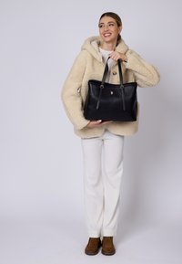 Borsa a tote in pelle nera con doppi manici, dotata di accessori in oro e di un piccolo emblema. Indossata da una figura in giacca in pile beige e pantaloni chiari.