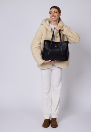 Sac en toile noir en cuir avec deux poignées, doté de quincaillerie dorée et d'un petit emblème. Porté par une silhouette vêtue d'une veste en polaire beige et d'un pantalon clair.