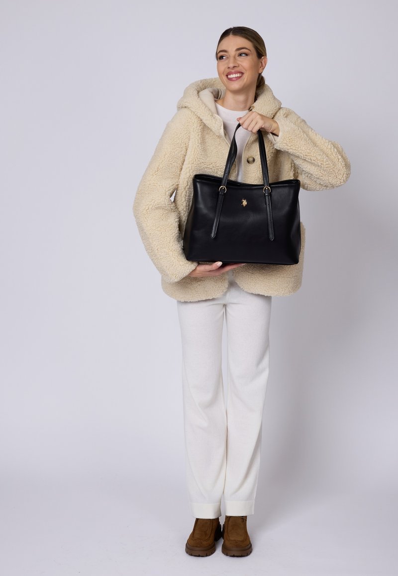 Borsa a tote in pelle nera con doppi manici, dotata di accessori in oro e di un piccolo emblema. Indossata da una figura in giacca in pile beige e pantaloni chiari.