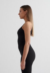 Črna oprijeta majica brez rokavov z tankimi naramnicami in brezšivno zasnovo, v kombinaciji s črnimi legicami z visokim pasom. Gladka tekstura, minimalni detajli.