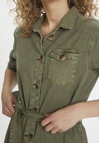 Olívově zelené džínové šaty košilového střihu s knoflíkovým zapínáním, náprsní kapsou a páskem v pase. Obsahují krátké rukávy a měkkou, texturovanou tkaninu.