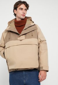 Veste matelassée beige avec capuche, dotée d'un zip côtelé à l'avant, de cordons de serrage réglables et d'accents texturés. Portée sur un col roulé couleur rouille.
