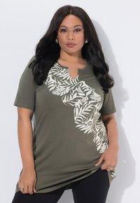 Frau mit langen schwarzen Haaren und Brille, die eine olivgrüne Tunika mit weißem Blätter-Muster trägt und vor einem schlichten hellen Hintergrund posiert.