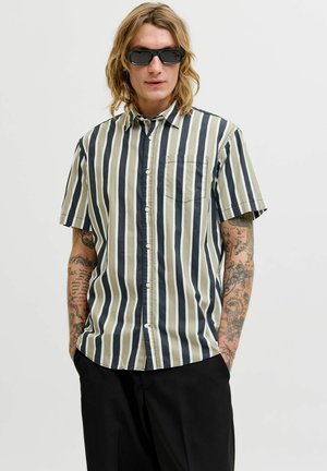 Mand med skulderlangt hår og tatoveringer iført sorte solbriller, en kortærmet skjorte med lodrette striber og sorte bukser, hænderne i lommerne.