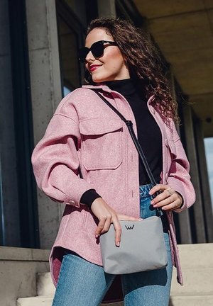 Rosa, wollartige Jacke mit großen Fronttaschen, kombiniert mit einem schwarzen Rollkragenpullover und blauen Jeans. Graue Tasche mit dezentem Logo-Detail.