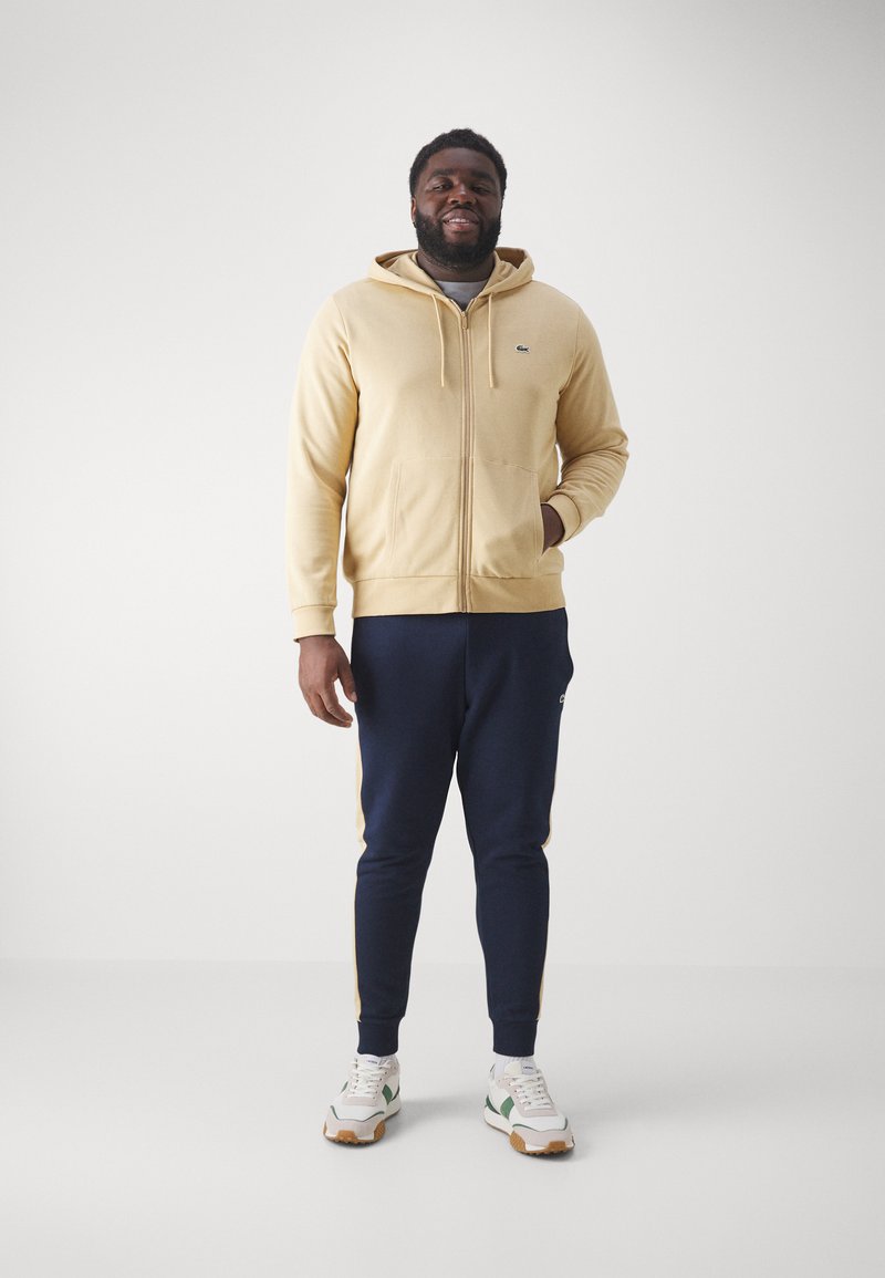 Beige zip-up hoodie met kangoeroezak, gecombineerd met marineblauwe joggers met zijstrepen. Witte sneakers met groene en bruine accenten.