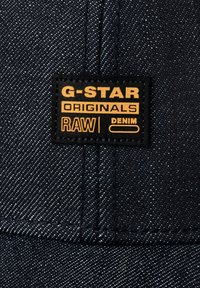 Mørkeblå denim caps med en teksturert finish. Den svarte merkelappen viser "G-STAR ORIGINALS RAW DENIM" i fet oransje skrifttype.