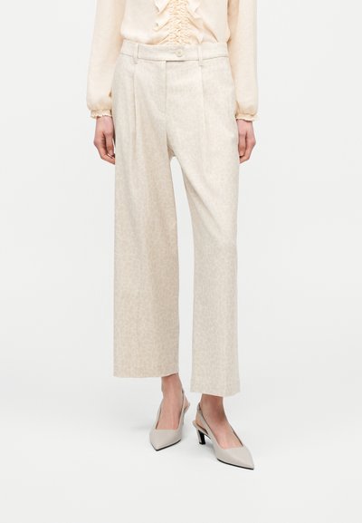 Femme portant un pantalon beige à taille haute et jambes larges avec un motif subtil, associé à un chemisier crème à volants et des chaussures à talons ouvertes beige.