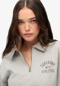 Sudadera con cremallera de color gris claro que presenta un cuello tipo mao, el texto "SUPERDRY ATHLETICS" y un gráfico de la bandera americana. Textura de tela suave.