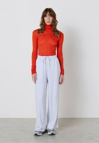 Sieviete ar viļņainiem brūniem matiem, kas ģērbusies sarkanā punktainā turtleneckā, gaiši pelēkās platu kāju sporta biksēs un pelēkos sporta apavos, stāv pretī baltai siena.