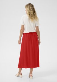 Jupe maxi rouge en tissu léger, dotée d'une taille froncée. Associée à un t-shirt blanc et des chaussures à talons blocs beiges.
