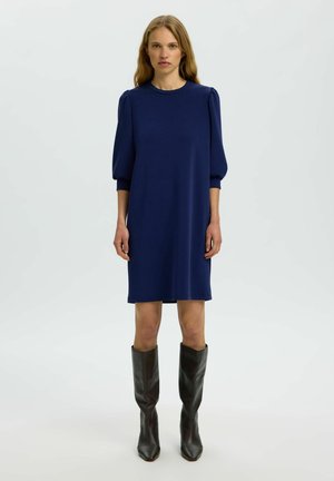 Frau in einem knielangen marineblauen Kleid mit gepufften Dreiviertelärmeln und schwarzen Lederstiefeln bis zur Mitte der Wade, steht vor weißem Hintergrund.
