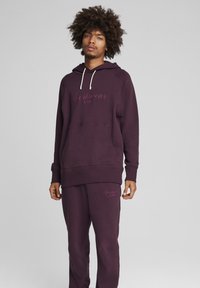 Lilla hættebluse med snører, frontlomme og matchende sweatpants; har tekstlogo i lysere lilla. Blødt, struktureret stof.