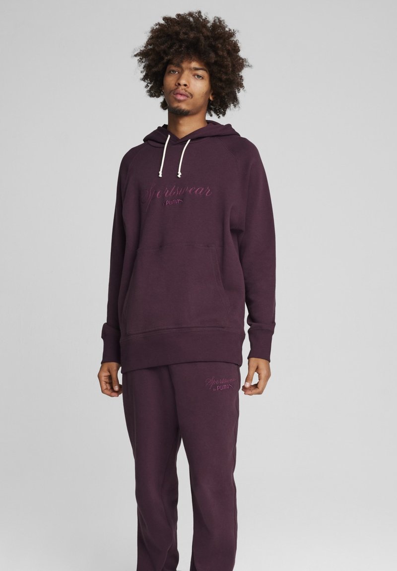 Lilla hættebluse med snører, frontlomme og matchende sweatpants; har tekstlogo i lysere lilla. Blødt, struktureret stof.
