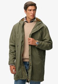Olive green parka with a zip and snap closure, featuring a fur-lined hood, side pockets, and a casual fit over a beige sweater and plaid shirt.
