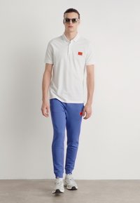 Baltas polo krekls ar mazu sarkanu logo uzšuvumu, apvienots ar pieguļošām zilām sporta biksēm un pelēkiem sporta apaviem. Modelis nēsā saulesbrilles.