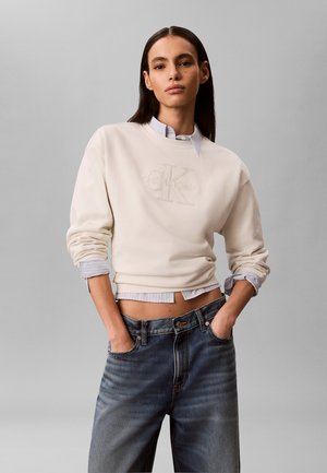 Młoda kobieta w białej bluzie Calvin Klein nałożonej na koszulę w paski, do tego luźne sprane dżinsy, ręce w kieszeniach.