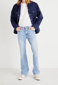 Jeans a zampa d'elefante in denim blu, lavaggio chiaro con leggeri effetti di scoloritura, abbinati a una t-shirt bianca a girocollo e a una giacca button-up blu navy, scarpe da ginnastica casual.