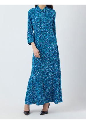 Femme portant une robe chemise longue fleurie bleue avec boutons, manches trois-quarts, et des escarpins noirs pointus.