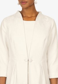 White textured jacket with a subtle floral pattern, soft collar, and two fabric-covered buttons at the waist, displayed over a matching top.